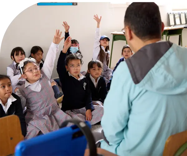 Foto de un salon de clase, se ve de espaldas un profesor y frente a el 8 niñas, algunas con sus abrazos arriba. 