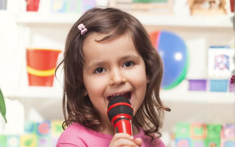 Niña pequeña cantando con un micrófono de juguete, rodeada de juguetes y cubos con letras